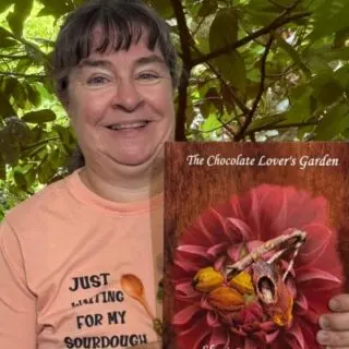 Sheri Ann Richerson smiling and holding her book The Chocolate Lover’s Garden in front of the lush green foliage of a Theobroma cacao tree in bloom.