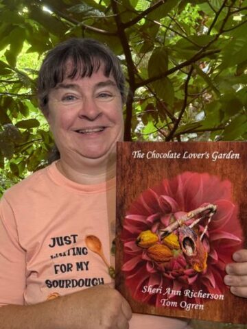 Sheri Ann Richerson smiling and holding her book The Chocolate Lover’s Garden in front of the lush green foliage of a Theobroma cacao tree in bloom.